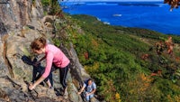 two people climbing up a rocky cliff with the ocean in the background