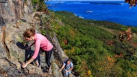 two people climbing up a rocky cliff with the ocean in the background